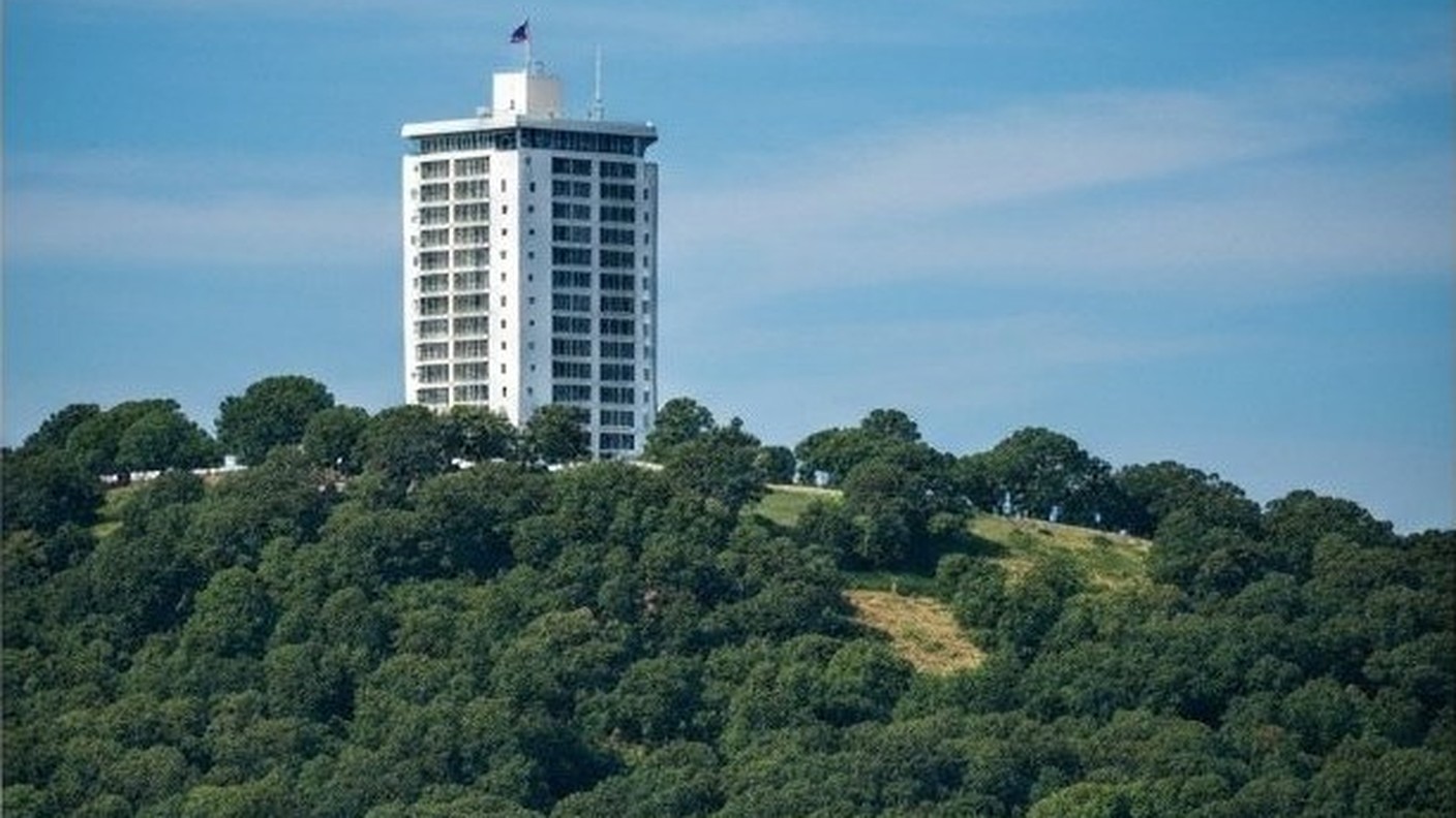tall white residential tower on a hill surrounded by green trees, blue sky